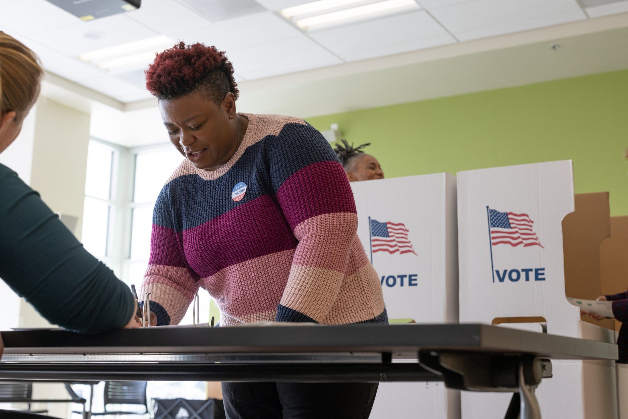 A voter wearing an "I Voted" sticker writes at a table in front of voting booths.
